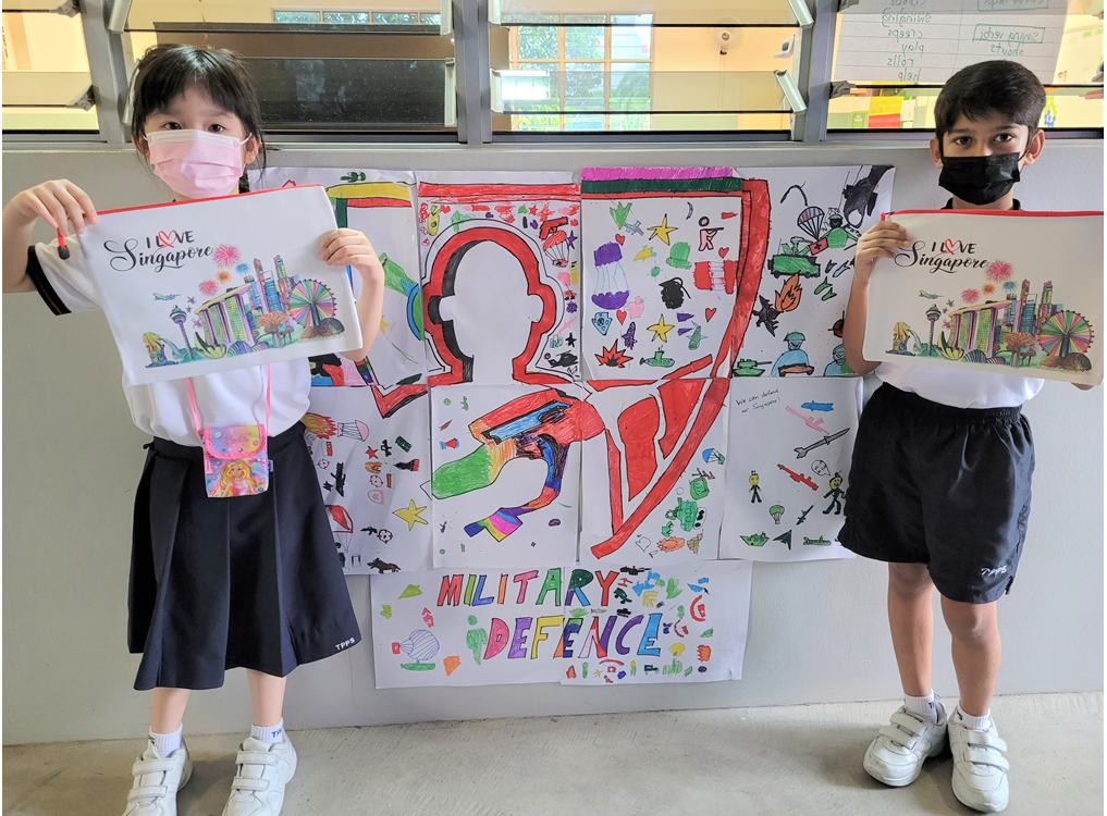 Two masked children hold Singapore-themed artwork in front of a colorful "Military Defence" poster on a wall.